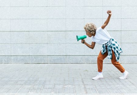 Angry African American woman yelling on megaphone while protesting about something outdoors on the street. Communication concept.