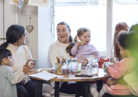 A medium shot of three toddlers and their mothers wearing casual clothing on a winter's day in Northumberland. They are at a preschool where they sit around a table, talk and enjoy a cup of tea and snacks.